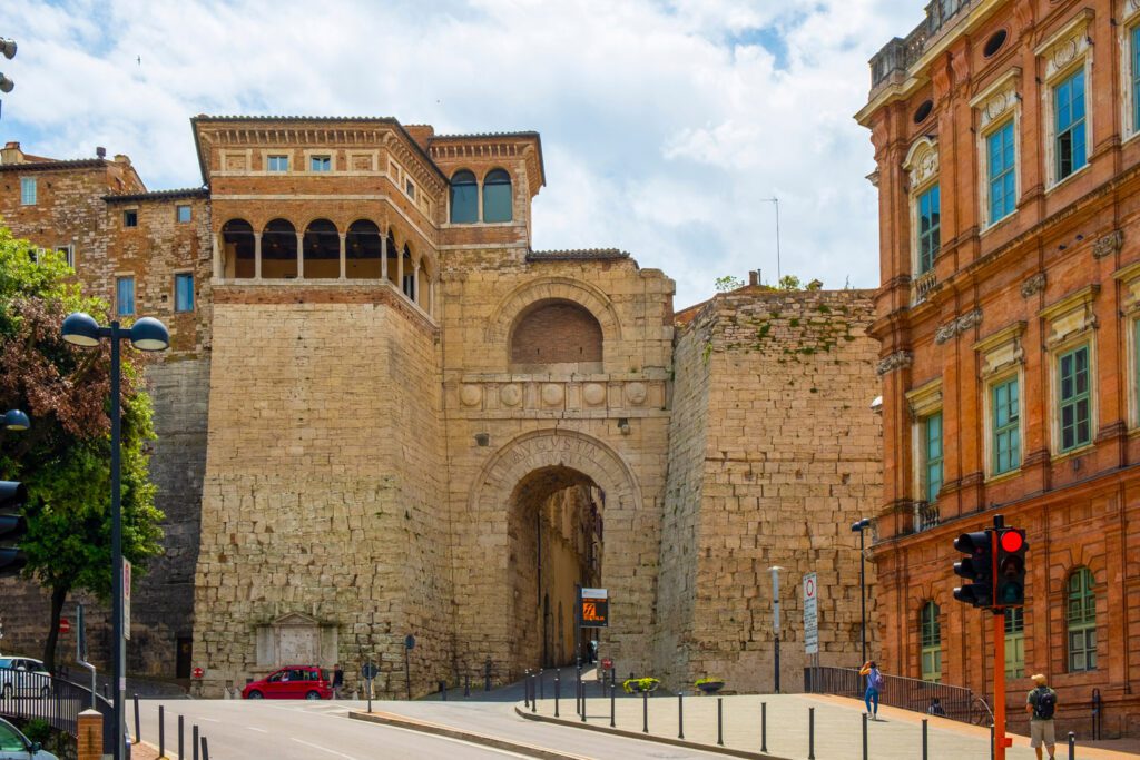 Vista frontale dell'Arco Etrusco. Sulla destra si scorge un palazzo rosso, l'università per Stranieri di Perugia. L'arco appare, imponente, direttamente affacciato sulla strada.