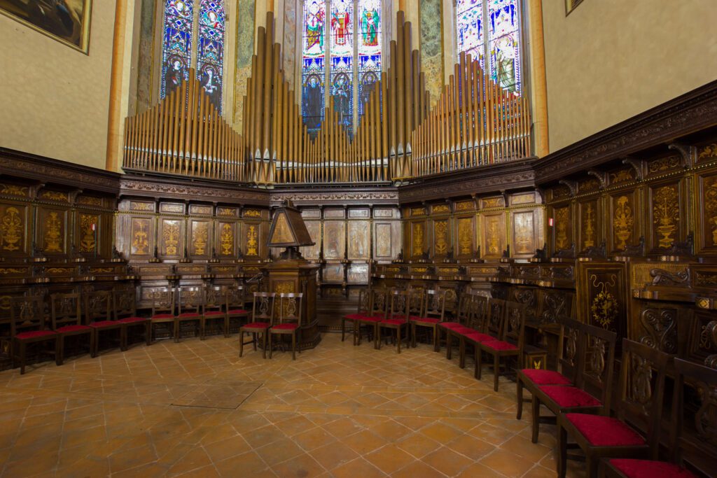 Vista del coro ligneo retrostante all'abside della cattedrale di San Lorenzo. Il coro ligneo, intagliato e intarsiato, è addossato all'abside e ne segue l'andamento semicircolare. Al di sopra di esso parte del grande organo della chiesa e le ampie vetrate dell'abside.