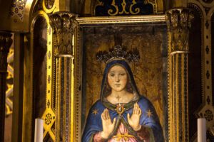 Dettaglio della Madonna delle Grazie nella cattedrale di San Lorenzo a Perugia. La vista della parte superiore dell’opera mostra la cornice dorata che contiene l’immagine di Maria. Le mani sono alzate poco sopra il pancione, in segno di accoglienza. Il volto è amorevole. Indossa un manto blu cn delle stelle dorate, e sopra il suo capo è presente una corona in pietre vere che sporge dalla superficie dell’opera.