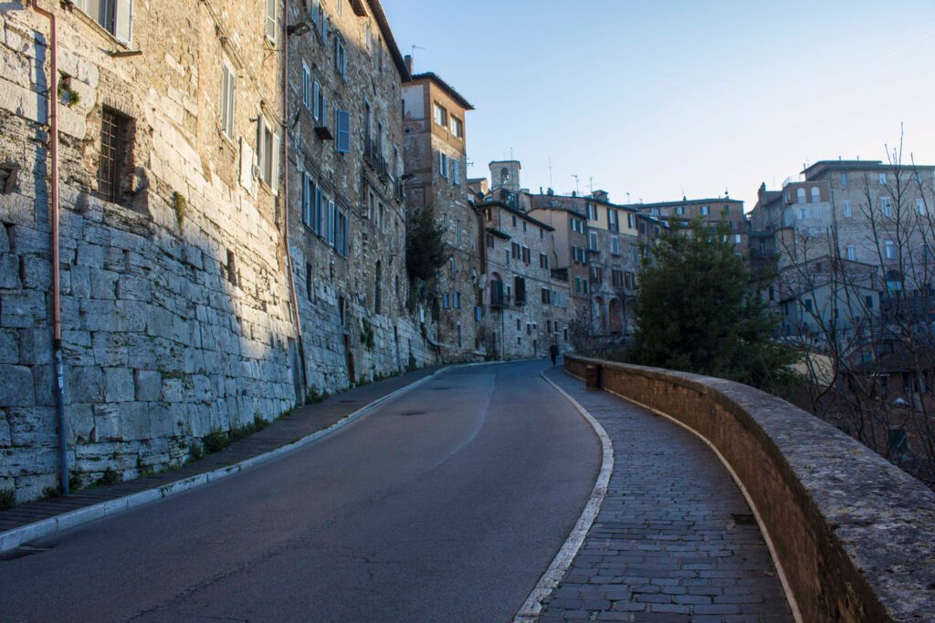 Vista della strada che si diparte a destra dell'arco etrusco. Sulla sinistra il cornicione del camminamento pedonale che permette una vista panoramica sulla zona universitaria e sulle scalette dell'acquedotto. A sinistra le mura etrusche che si fondono a quelle dell'abitato.