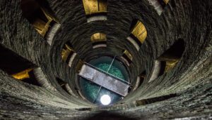 Sguardo dall’alto verso il fondo del pozzo. Sulle mura si vedono alcune finestre mentre sul fondo il ponte in ferro sospeso sull’acqua.