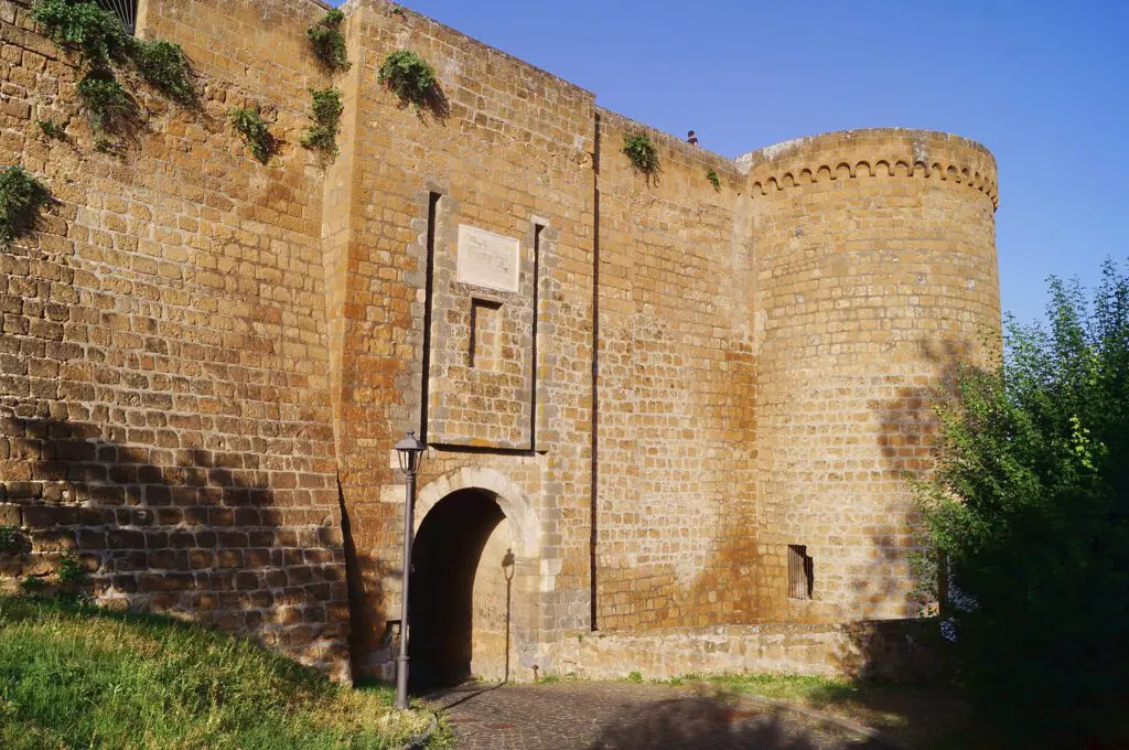 Vista leggermente scorciata dell’ingreso alla fortezza Albornoz. Su un alto muro di mattoni, che termina sulla destra con un torrione circolare, si apre il portone di accesso alle mura.