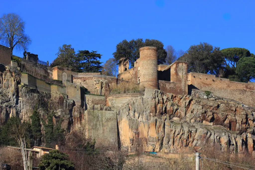 Vista della Rocca Albornoz dai piedi della Rupe. Le mura si intrecciano con la parete rocciosa e la costeggiano, e il torrione circolare posa al limite dello strapiombo della Rupe.