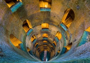 vista interna dall’alto verso il basso del Pozzo di San Patrizio a Orvieto. Le pareti interne sono interrotte regolarmente da ampie aperture da cui si intravedono le scale a chiocciola interne che scendono verso il fondo del pozzo. Sul fondo del pozzo, dei turisti stanno attraversando il piccolo ponte che collega le due scalinate interne, una che scende verso il basso e una che risale verso l’alto