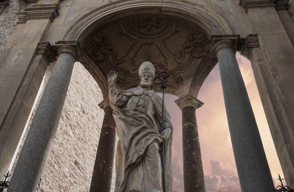 Vista leggermente dal basso di una statua di Sant'Ubaldo a Gubbio. La statua, in pietra, rappresenta Sant'Ubaldo con le vesti da vescovo e si trova all'interno di un baldacchino in pietra.