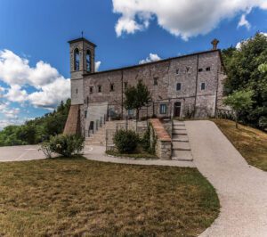 Vista scorciata dell'esterno della Basilica di Sant'Ubaldo. In primo piano un'aiuola attraversata da una strada che porta all'accesso attraverso una scalinata o una piccola salita senza barriere architettoniche.