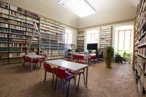 Vista di una delle sale della biblioteca di Palazzo della Corgna. Le pareti sono ricoperte da scaffali pieni di libri, tranne che in tre punti dove finestrini fanno filtrare grandi quantità di luce. Al centro della stanza alcuni tavoli con sedie per consultare comodamente i testi.