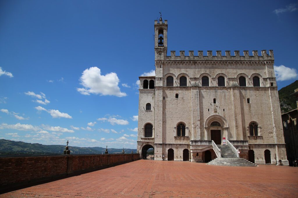 Vista della facciata di Palazzo dei Consoli, sulla sinistra il delimitare di Piazza Grande lascia intravedere il paesaggio delle campagne umbre.