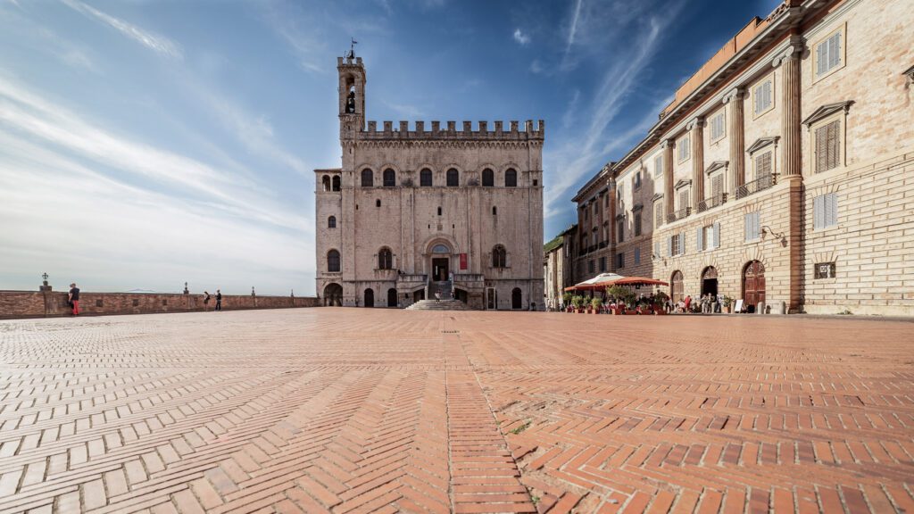 Vista della facciata di Palazzo dei Consoli dalla parte più lontana della piazza, il cui pavimento rosso si specchia sulla pietra bianca del palazzo.