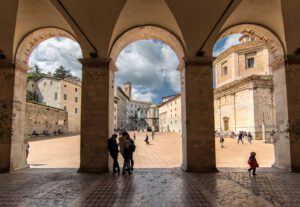 Vista della piazza antistante lil Duomo di Spoleto dall'ingresso della chiesa. Gli archi dividono scenicamente la vista sulla piazza svelando l'attività di alcune persone che passeggiano.