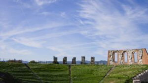 Vista della cavea e di alcune mura del Teatro Romano sovrastato da un grande cielo azzurro.