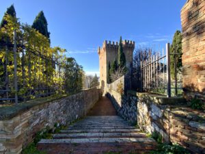 Vista laterale del cassero di Porta Sant'Angelo. In primo piano la stretta via, delimitata ai lati da bassi muretti e inferriate, che unisce l'ingresso della Torre del cassero con lo spazio antistante al tempietto, cioè la chiesa di San Michele Arcangelo. La vista della via porta al cassero, che si staglia con i suoi merli nel cielo azzurro.