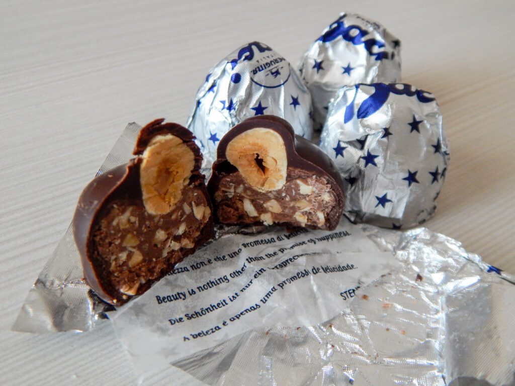 Vista di Baci Perugina. In primo piano un bacio perugino aperto a metà all'interno del quale si vede la nocciola intera e il ripieno di cioccolata. Sotto le due metà del cioccolatino è steso l'inolucro, formato da un biglietto con citazioni celebri su amore e amicizia e il classico rivestimento in alluminio argentato con stelle blu. Sullo sfondo altri due baci perugina, incartati.