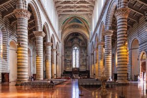 Interno del Duomo di Orvieto con vista dalla navata centrale rivolta verso l’altare. Gli spazi sono ampi, divisi in tre navate dalle colonne di marmo bicromo. La luce filtra dalle finestre in alabastro.