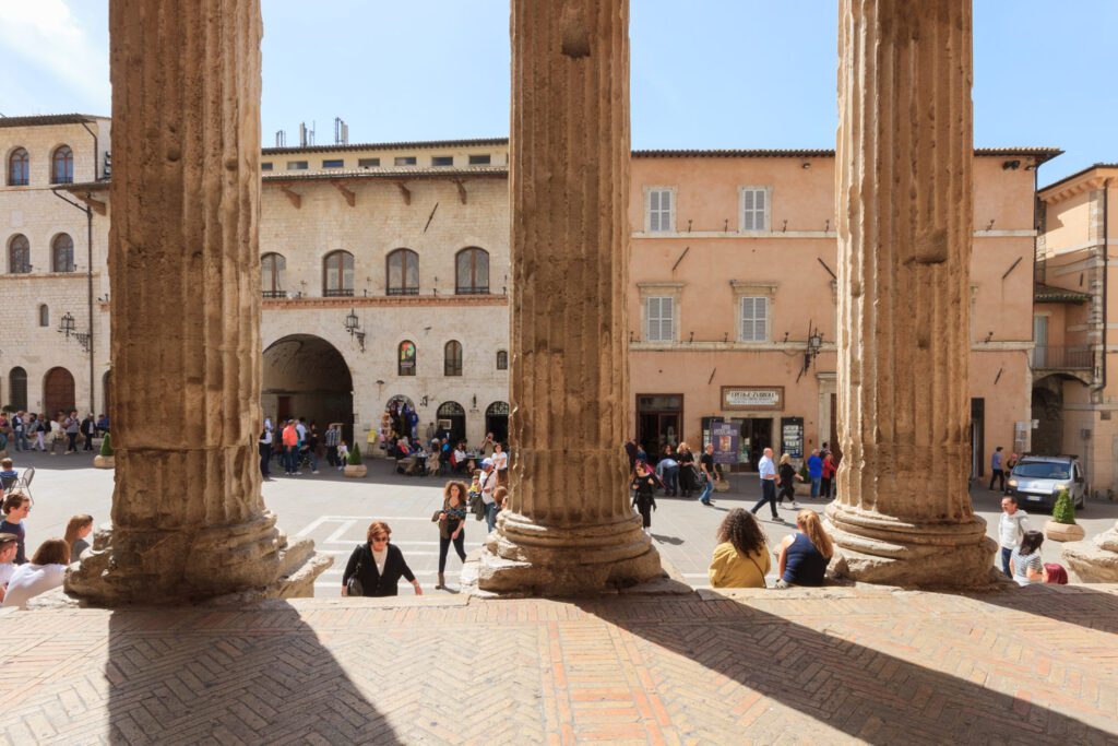 Vista di Piazza del Comune dal pronao del tempio di Minerva. Le colonne nascondono parti della piazza per poi svelare l'attività dei turisti in piazza.