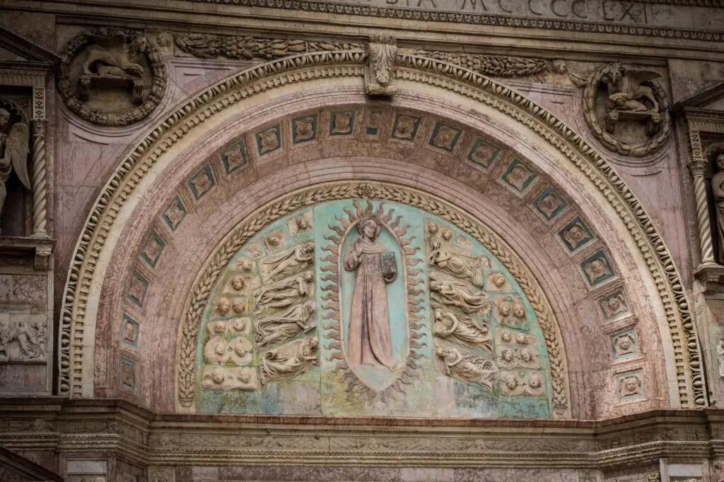 Dettaglio della facciata della chiesa di San Bernardino a Perugia. Sopra il portale di ingresso, all'interno di una cornice semicircolare rivolta verso il basso, spicca la scultura di San Bernardino, realizzata in pietra rossa, come la facciata, e con sfondo turchese.