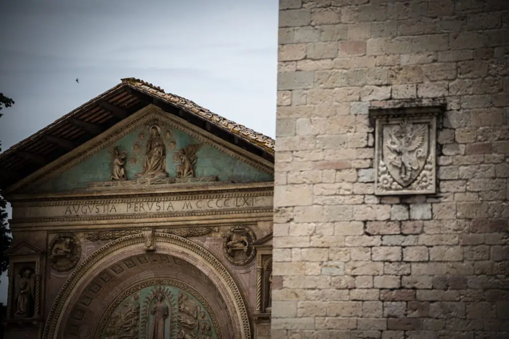 Vista scorciata di un dettaglio sulla facciata della chiesa di San Francesco al Prato. Sull'estrema sinistra della facciata si trova una decorazione scultorea che riporta il grifo, simbolo di perugia. Alla sinistra del muro, compare la parte superiore della facciata di San Bernardino, colta prospetticamente.