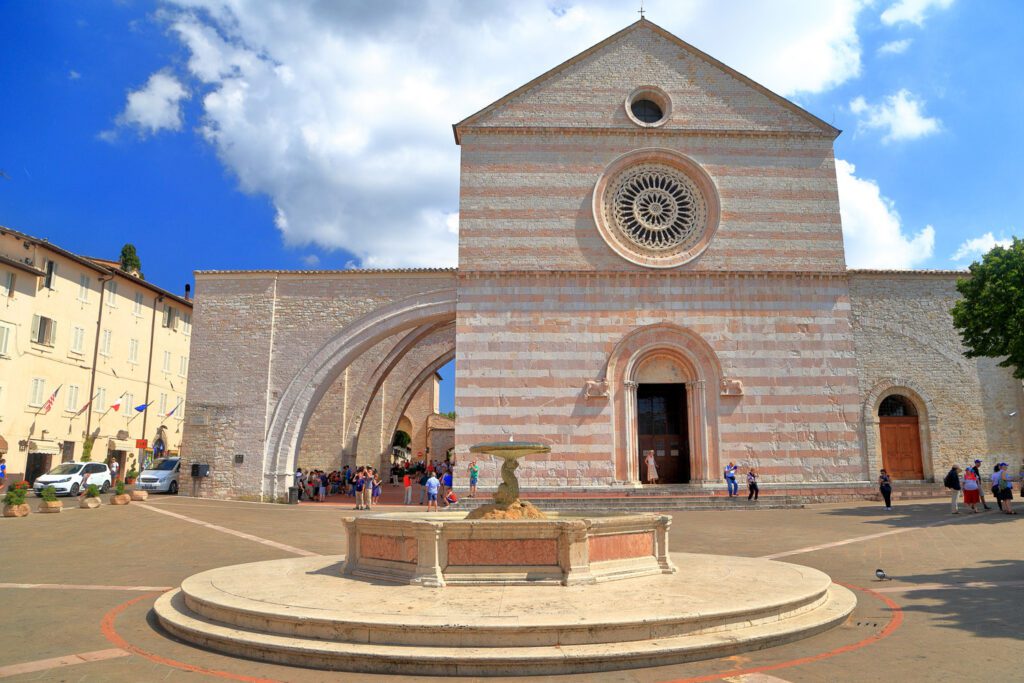 Vista frontale della chiesa di Santa Chiara. In primo piano la piccola fontana nella piazza antistante. Sullo sfondo la chiesa, con i suoi arconi sulla sinistra.
