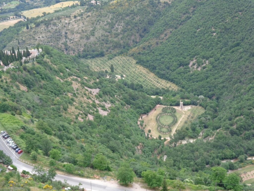 immagine panoramica del Bosco di San Francesco e del territorio montuoso circostante dall’alto, in cui si distingue l’opera di land art fatta di 3 cerchi concentrici Terzo Paradiso di Michelangelo Pistoletto, a fianco della quale svetta una torre d’osservazione. A margine, in basso sulla sinistra, la strada che costeggia il bosco con delle auto parcheggiate.