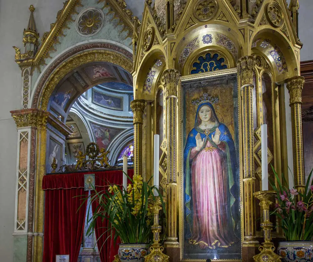 Il dipinto della Madonna delle Grazie del Perugino visto per intero, racchiuso all’interno di un tabernacolo rivestito d’oro con vetri, affiancato da ceri e composizioni floreali. Sullo sfondo una parete della Cattedrale di San Lorenzo con un panneggio che conduce a una cappella affrescata e con dettagli dorati.
