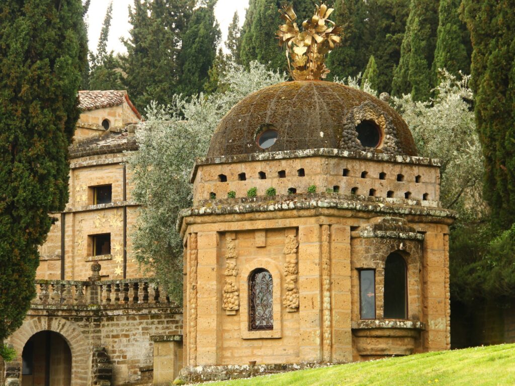 Exterior view of a sacred chapel located in the gardens of the Scarzuola. It is surrounded by trees, while other buildings in the park can be glimpsed in the background.