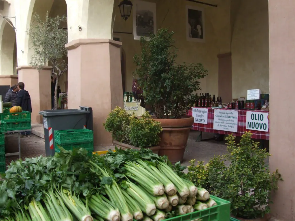 Photo taken on the day of the farmers' market in Trevi. The market takes place inside a courtyard bordered by a portico. In the foreground, inside the courtyard, there are bunches of celery lying one on top of the other inside a large plastic basket. Other plastic baskets indicate that a market is taking place and there are two people on the left who are talking. In the background, under the arcade, there are many bottles of oil displayed for sale on long table. Next to the columns of the arcade, there are large plants in a pot used as decorative elements.