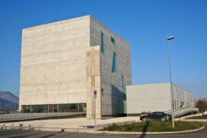 vista esterna della chiesa di San Paolo a Foligno e dell’area circostante, progettate dagli architetti Massimiliano e Doriana Fuksas. Sulla parte sinistra della foto c’è l’edificio più grande, dove all’interno è collocata la chiesa. Da un lato la facciata principale della chiesa è fatta in cemento armato, senza aperture; una passerella leggermene in salita conduce alla base dell’edificio, dove una lunga vetrata costituisce l’ingresso della chiesa. Sulla facciata laterale invece ci sono delle finestre di dimensioni e forme diverse. Di fronte alla chiesa, sulla destra dell’immagine, si trova un altro edificio in cemento armato più piccolo, con delle finestre disposte su due file lungo il lato lungo, rivolve verso l’esterno. In un piccolo piazzale accanto alla passerella d’ingresso, al centro dell’immagine, si trova un crocifisso scolpito in un blocco di cemento e marmo bianco di Carrara alto 13 metri, realizzato dall’artista Enzo Cucchi. Di fronte alla chiesa, c’è un parcheggio in cui sono parcheggiate due auto.