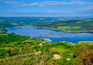 foto panoramica dall’alto del Lago di Corbara. Al centro dell’immagine si trova una parte del lago situato all’interno del Parco Fluviale del Fiume Tevere. Tutto intorno è circondato da bassi rilievi ricoperti di vegetazione. Sparse nella parte bassa della foto, si intravedono i tetti di alcune abitazioni tra i boschi.