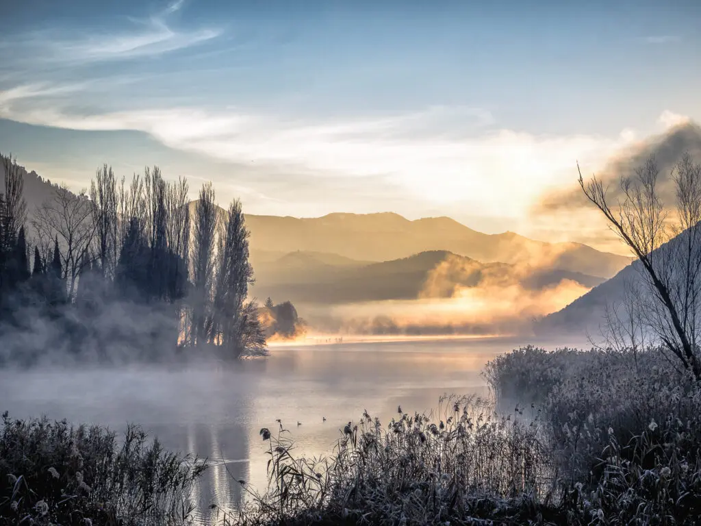 Foto del Lago di Piediluco scattata alle prime luci dell’alba. Il paesaggio lacustre, circondato dalle montagne, è ricoperto di nebbia e una leggera luce del mattino si riflette con diverse sfumature sulla nebbia.