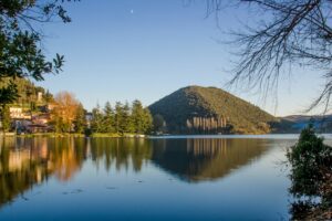 Vista del Lago di Piediluco e delle montagne che lo circondano. Sulla riva a sinistra dell’immagine, un gruppo di case è illuminato dai raggi del sole e accanto si trova un piccolo gruppo di pini.