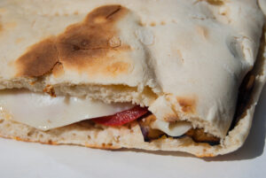 Close-up frontal shot of the hands of a lady holding the stretched out dough of torta al testo on the side edges. The dough rests on a wooden cutting board and has the typical shape of torta al testo, round and flattened.